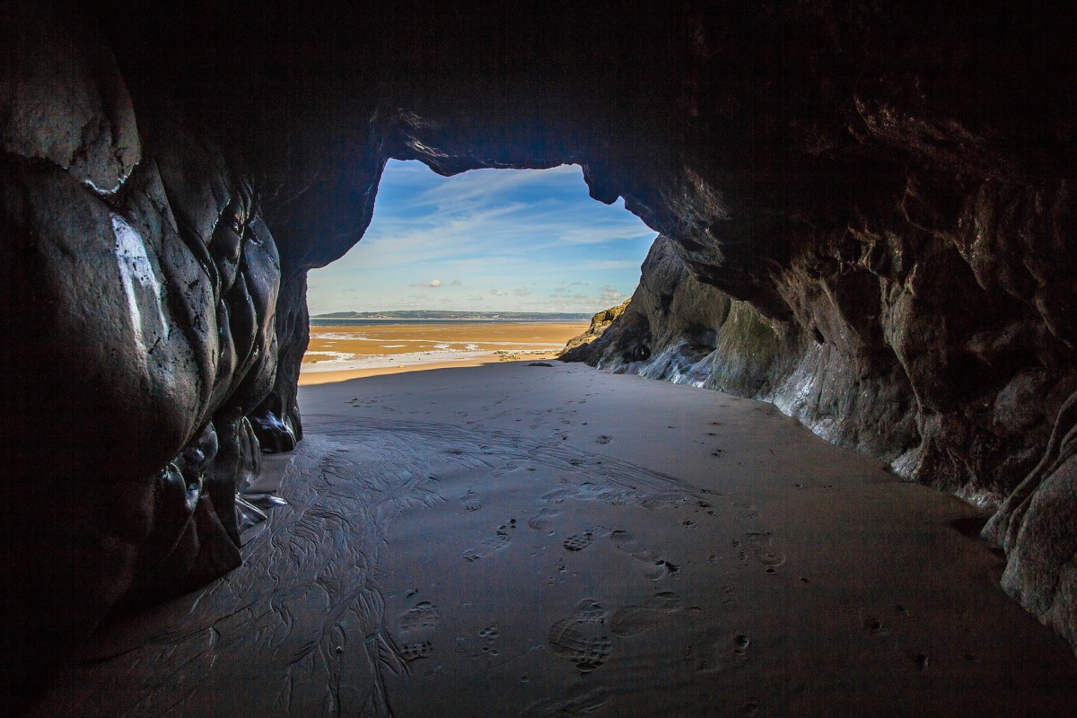 View out towards a beach at low tide, with a blue sky, from inside a sandy cave.