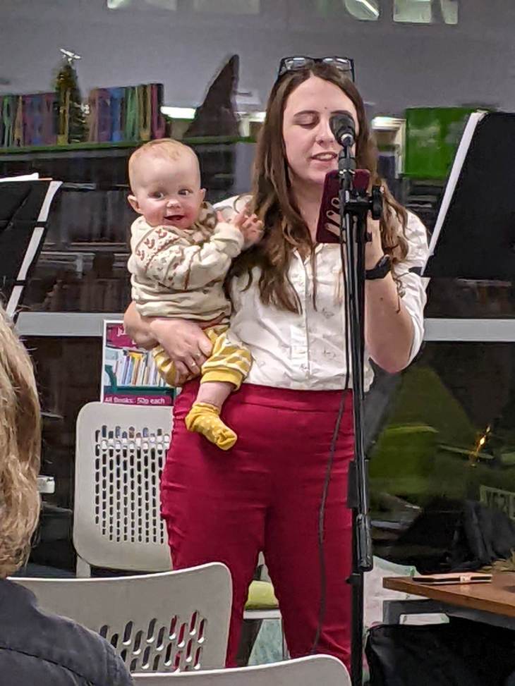 Me reading poetry at Crewe library in red trousers, with a white shirt with my daughter on my hip, dressed in yellow socks, yellow trousers, and a knitted beige jumper decorated with foxes. 